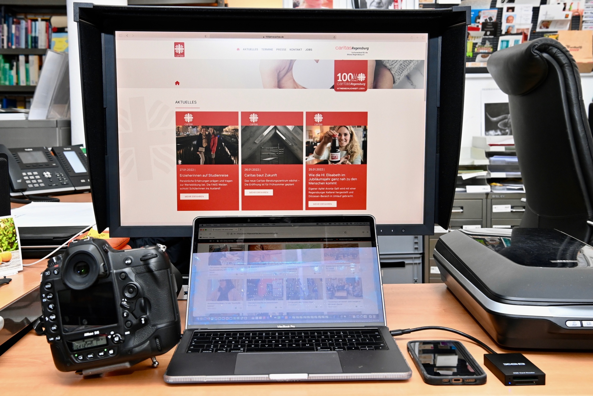 Kamera, Laptop und Monitor mit Website auf Schreibtisch.