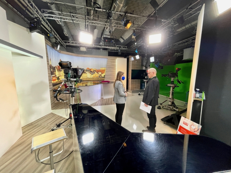 Ein modernes Fernsehstudio mit gebogener Theke und gro&szlig;em Bildschirm im Hintergrund. Der Raum ist hell ausgeleuchtet.