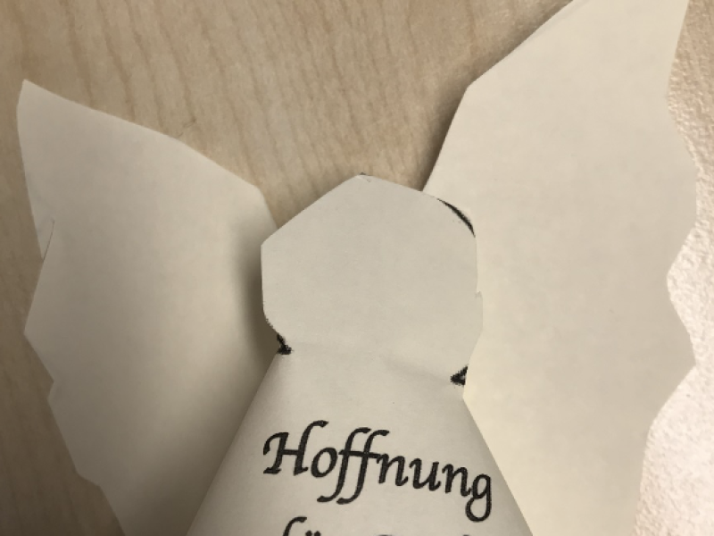 Ein gefaltetes wei&szlig;es Papierobjekt mit handschriftlichem Text liegt auf einem Tisch. Es erinnert an einen pers&ouml;nlichen Gru&szlig;.