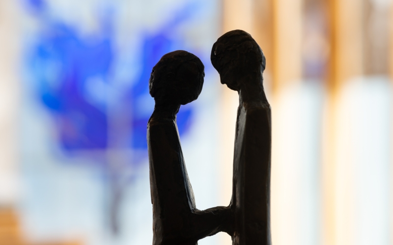Silhouette zweier Personen, die sich zugewandt gegenüberstehen und sich an den Händen halten. Im unscharfen Hintergrund sind blaue und wärmere Farbtöne erkennbar.