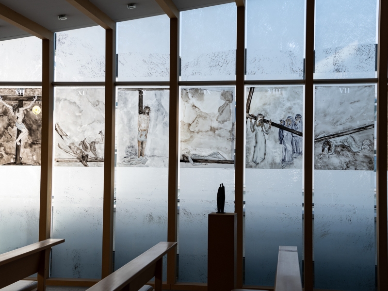 Blick durch gro&szlig;e Fenster mit Holzrahmen auf verschneite Berglandschaft; an der Fensterbank steht eine dunkle Skulptur, dahinter h&auml;ngen Kunstdrucke mit fig&uuml;rlichen Darstellungen.