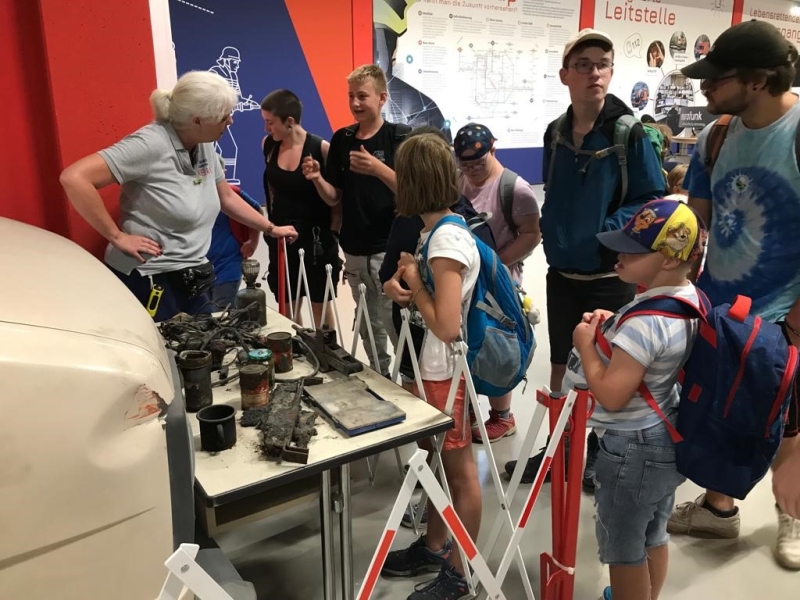 An einer Ausstellung stehen Besucher verschiedenen Alters um einen &auml;lteren Mann herum, der technische Ger&auml;te auf einem Tisch pr&auml;sentiert. Kinder und Erwachsene beobachten interessiert die Demonstration in einem Museum oder Ausstellungsraum mit roten und blauen W&auml;nden.