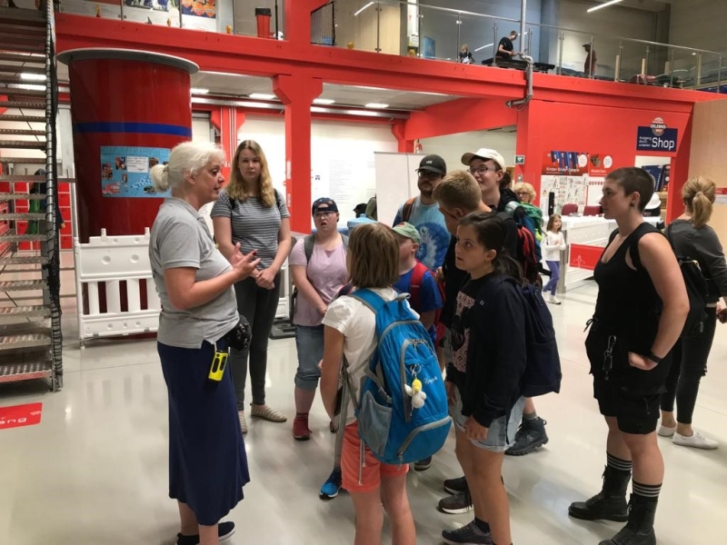 An einer Ausstellung stehen Besucher verschiedenen Alters um einen &auml;lteren Mann herum, der technische Ger&auml;te auf einem Tisch pr&auml;sentiert. Kinder und Erwachsene beobachten interessiert die Demonstration in einem Museum oder Ausstellungsraum mit roten und blauen W&auml;nden.