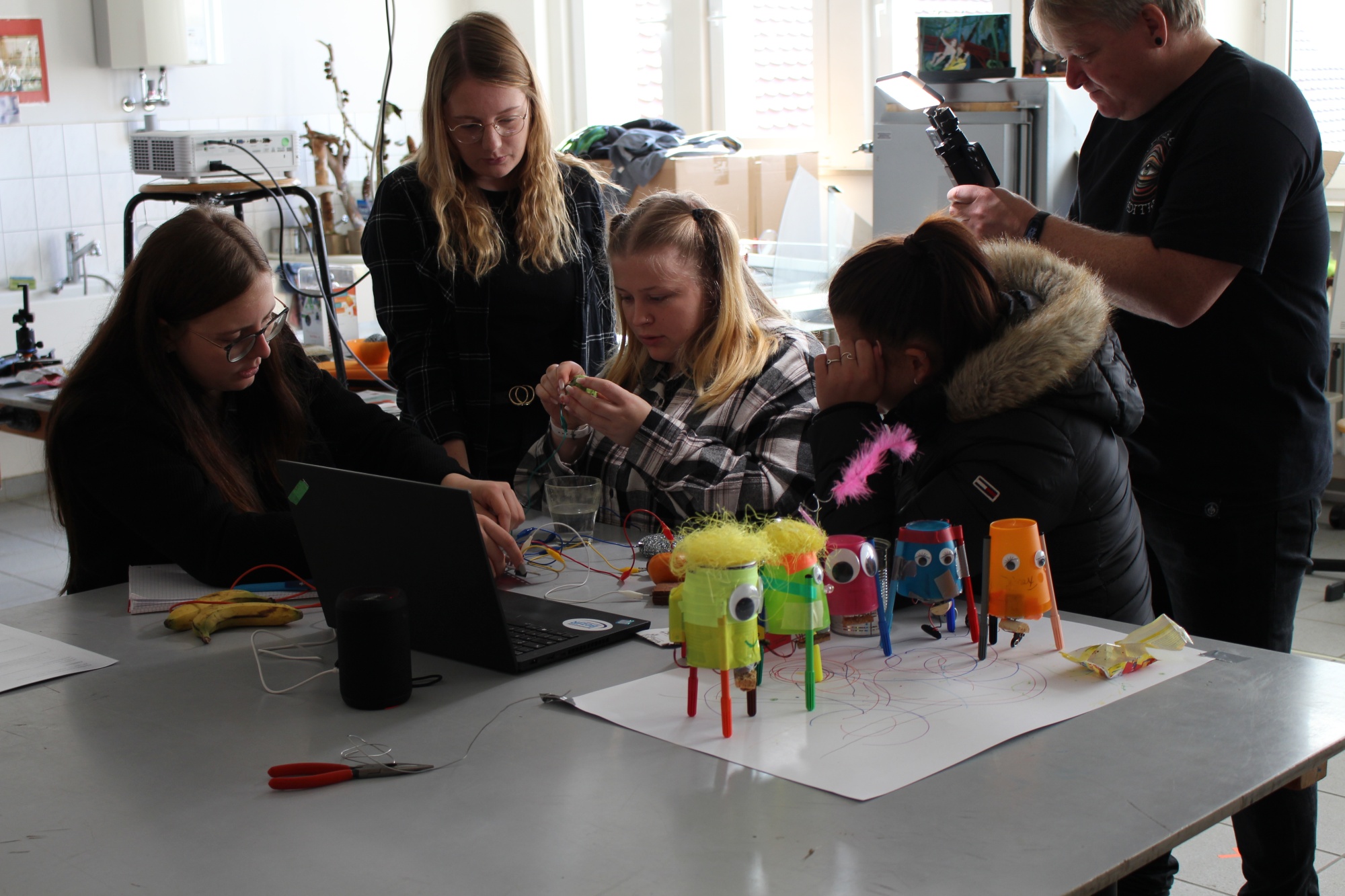 Fünf Personen unterschiedlichen Alters arbeiten gemeinsam an einem Tisch an bunten, selbstgebauten Objekten aus Recyclingmaterialien. Die Gruppe konzentriert sich auf ihre kreativen Handwerksprojekte in einem hellen Raum.