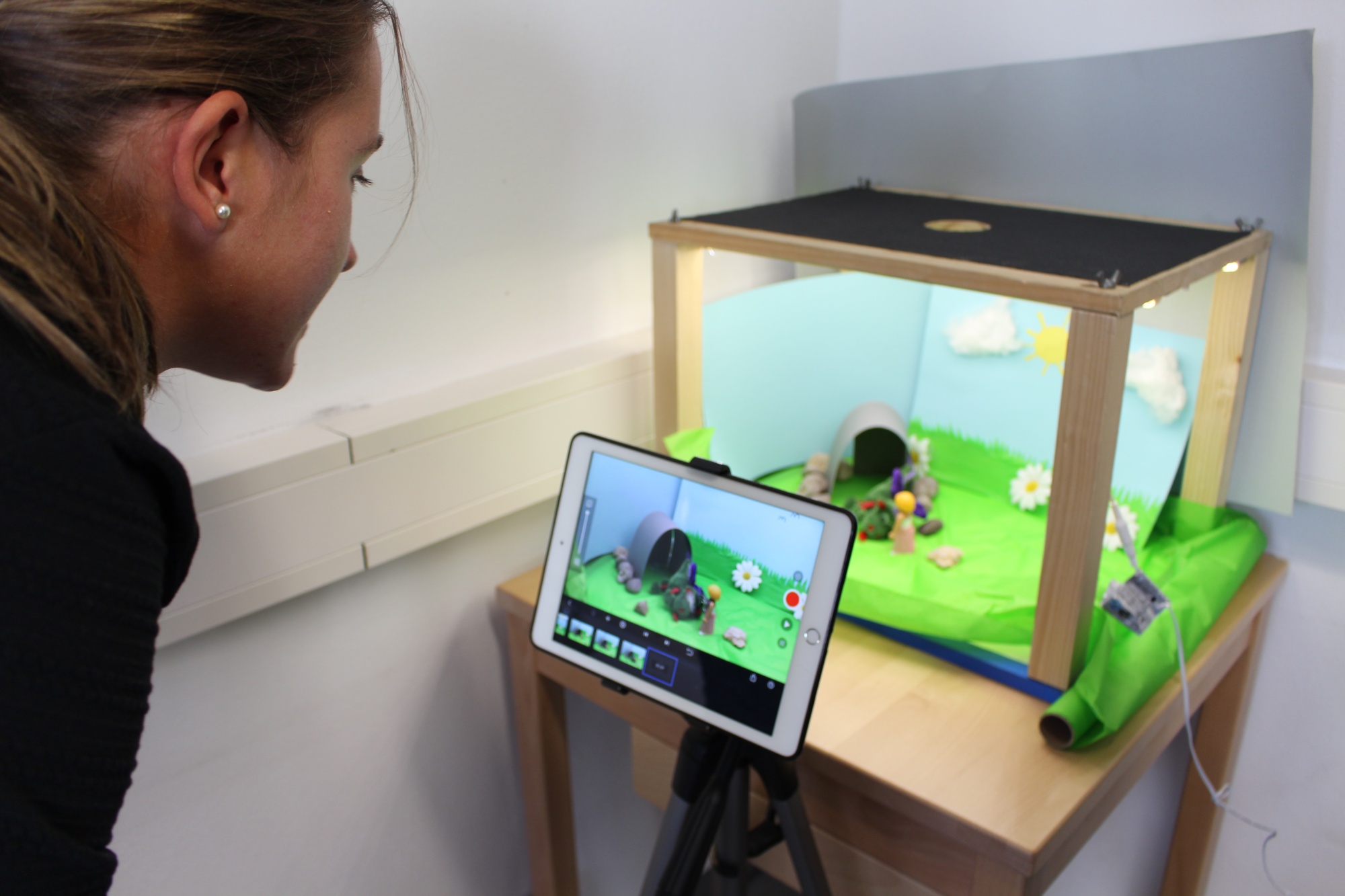 Eine Frau blickt auf ein interaktives Display mit grüner Landschaft und Figuren, während auf einem Tablet vor ihr die gleiche Szene zu sehen ist, die es ihr ermöglicht, die Ausstellung digital zu steuern.