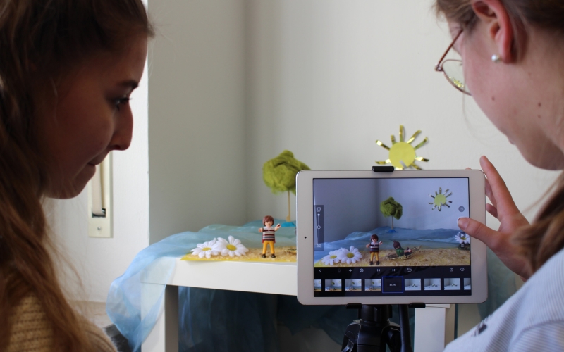 Eine Person hält ein Tablet in die Kamera, das einen Stop-Motion-Film mit einer Sandburg und Sonne zeigt, während zwei Personen das Ergebnis auf dem Bildschirm betrachten.