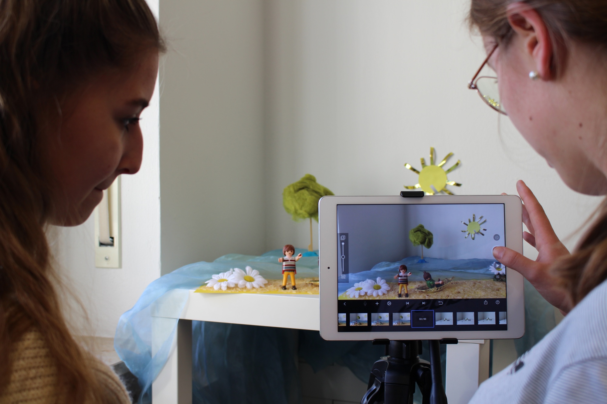 Eine Person hält ein Tablet in die Kamera, das einen Stop-Motion-Film mit einer Sandburg und Sonne zeigt, während zwei Personen das Ergebnis auf dem Bildschirm betrachten.